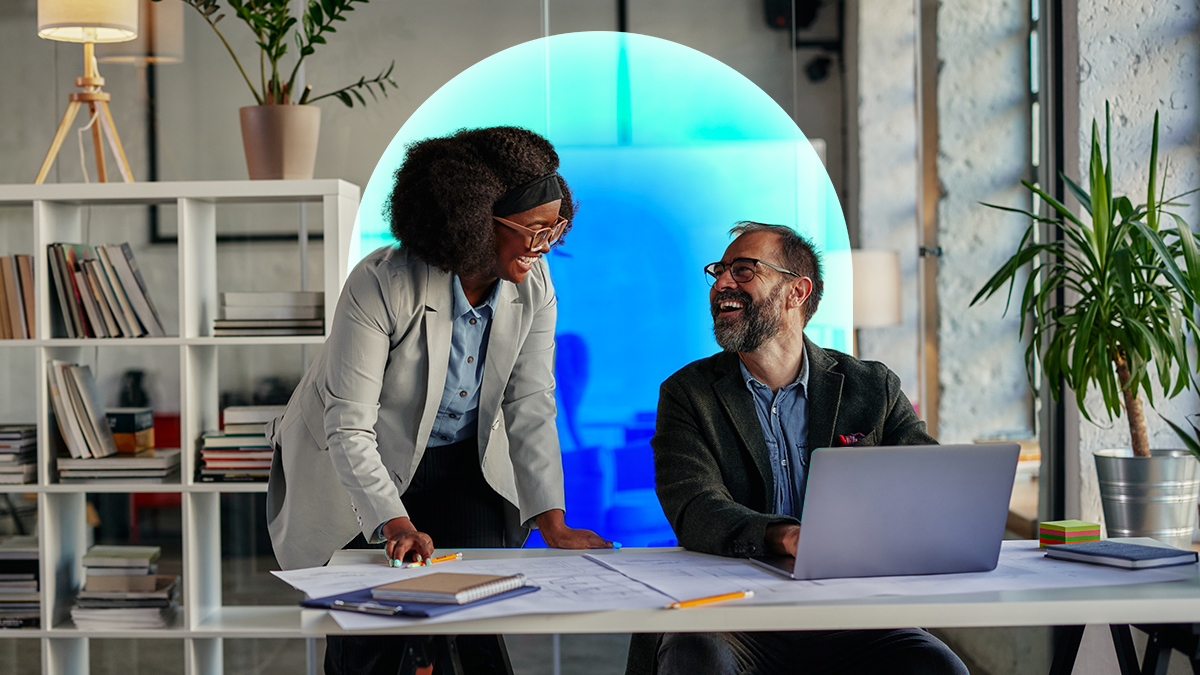 Corporate woman standing next to a corporate man on a laptop laughing